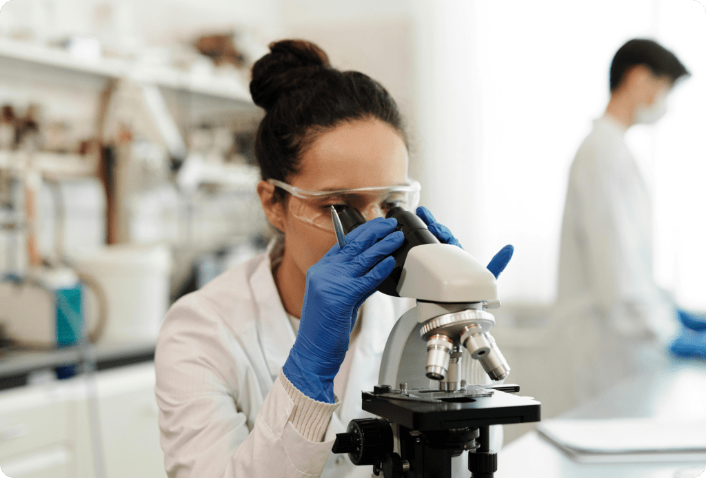 A laboratory scientist is examining a sample through a microscope while another researcher works in the background, representing medical research and diagnostic testing.