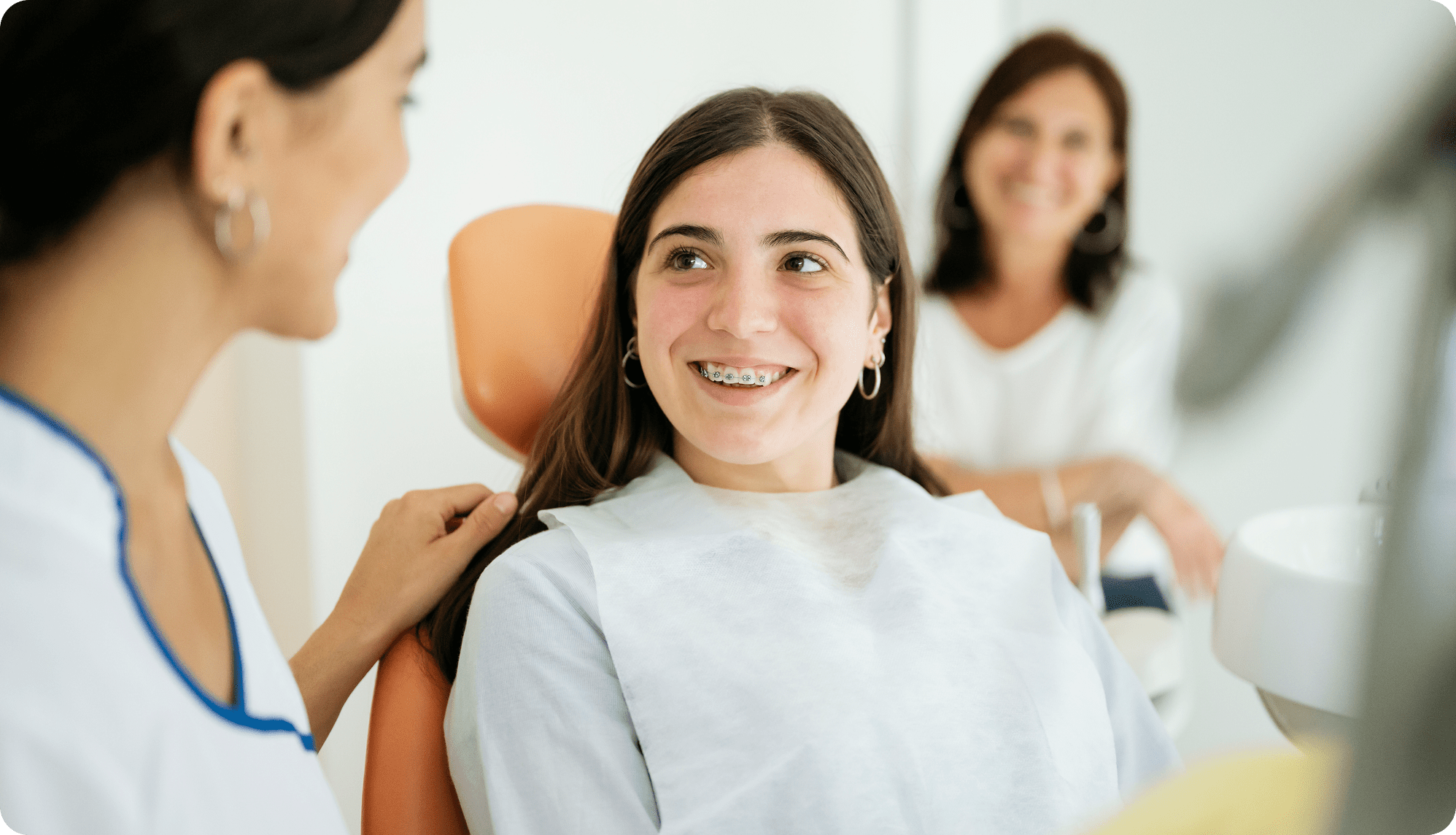 Teenage patient with braces smiling while talking with a dentist during a routine orthodontic checkup as a parent watches in the background.